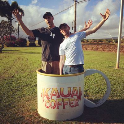 Daniel Robert and David Kennedy in a giant coffee cup, holding their arms up, in Hawaii that says Kauai Coffee.