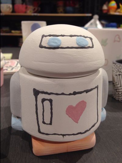 A small ceramic robot, painted with a red heart on its chest and blue eyes, ready to be fired.